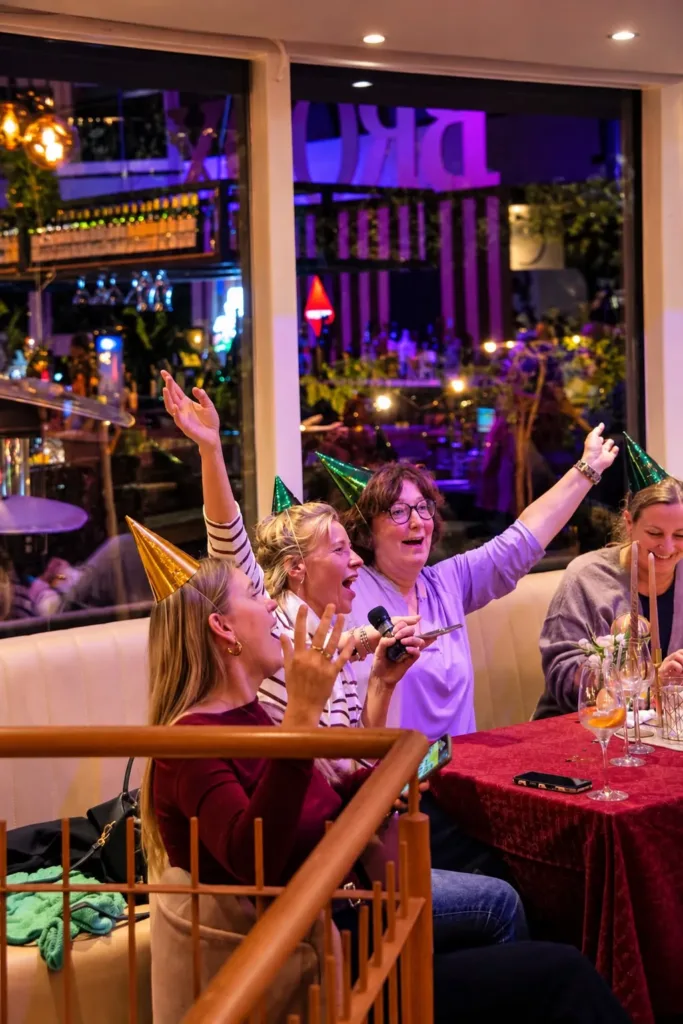 Vrouwen met feesthoedjes zingen en juichen met een microfoon aan een rode tafel tijdens een kerstborrel bij Broox Rotterdam